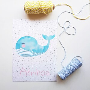 May include: A watercolor print of a blue whale with closed eyes and a pink mouth. The whale is swimming on a white background with gray dots. The name "Ainhoa" is written in pink below the whale.