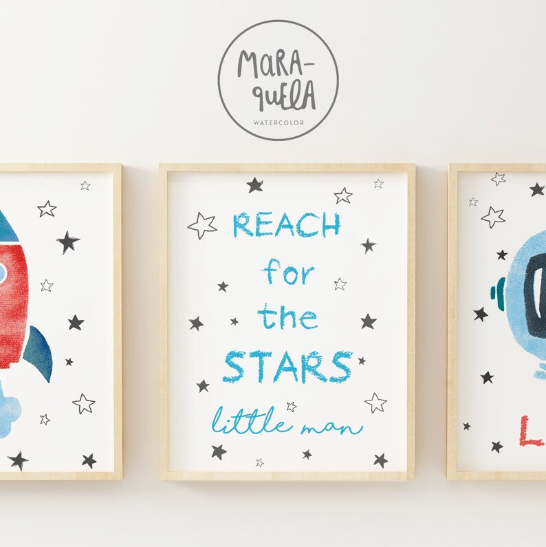 Pu&ograve; includere: Tre stampe incorniciate a tema spaziale. La stampa centrale presenta il testo "REACH for the STARS little man" in lettere blu e nere con stelle nere sparse intorno al testo. La stampa a sinistra presenta un razzo rosso e blu con stelle nere. La stampa a destra presenta un casco da astronauta blu con stelle nere.