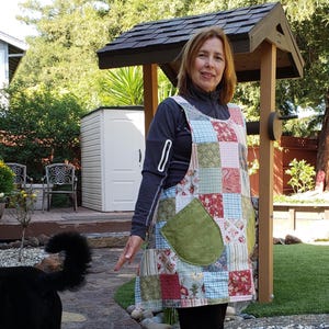 May include: A patchwork apron with a large green pocket is worn over a dark long-sleeved shirt and black pants. The apron features squares of various floral and patterned fabrics. A black dog stands on a brick patio.