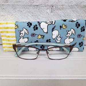 May include: Eyeglasses with a brown frame and light blue accents rest on a light blue case patterned with white cartoon dogs and black paw prints. A yellow and white striped case is to the left.