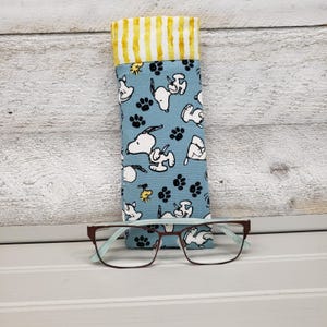 K&ouml;nnte beinhalten: Ein blaues Stoff-Brillenetui mit Snoopy-Muster und gelb-wei&szlig; gestreiftem Oberteil. Eine braune und hellblaue Brille liegt vor dem Etui. Das Etui hat ein sich wiederholendes Muster von Snoopy und Pfotenabdr&uuml;cken.