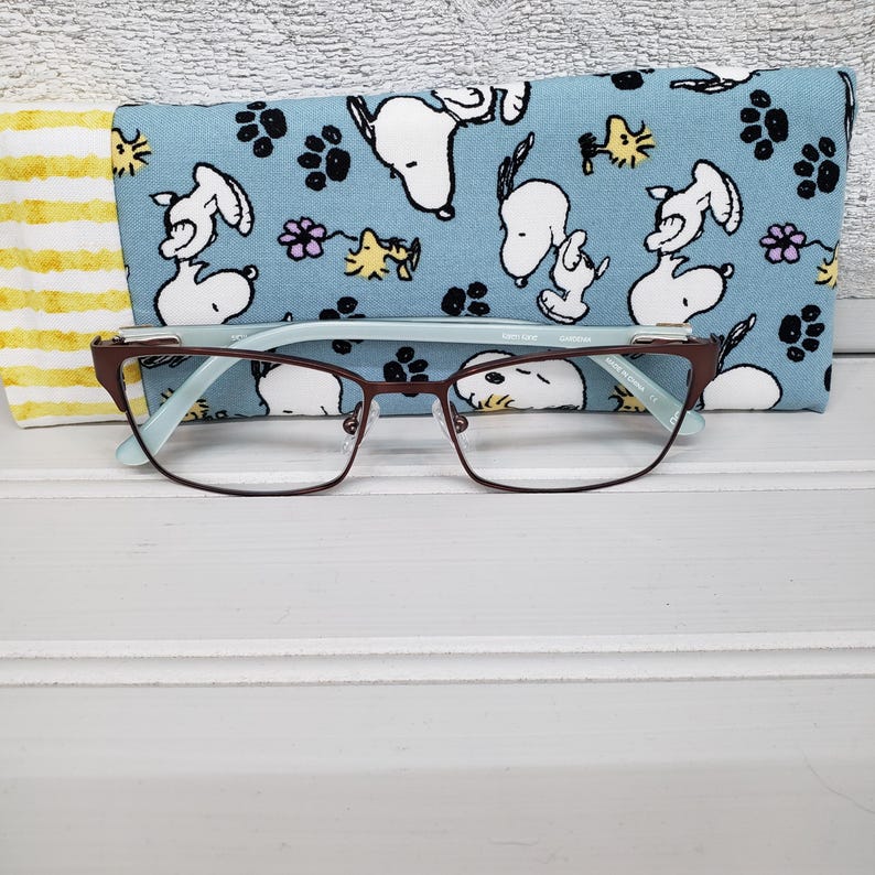 K&ouml;nnte beinhalten: Braune rechteckige Brille mit hellblauem Innenraum und einem blauen Etui mit Snoopy-Muster. Auf der linken Seite des Bildes ist ein gelb-wei&szlig; gestreifter Stoff zu sehen. Die Brille liegt auf einer wei&szlig;en Oberfl&auml;che.