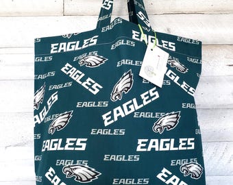 May include: A green tote bag with white text and a logo that says "EAGLES". The bag has a repeating pattern of the word "EAGLES" and a stylized eagle head. The bag is hanging from a metal hook.