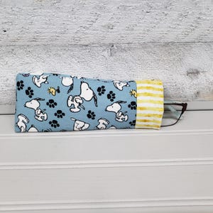 K&ouml;nnte beinhalten: Ein blaues Brillenetui aus Stoff mit Snoopy und Pfotenabdr&uuml;cken. Das Etui hat ein gelb-wei&szlig; gestreiftes Ende. Eine Brille ist teilweise im Inneren des Etuis zu sehen.
