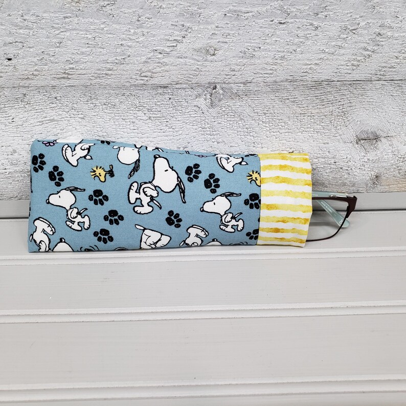 K&ouml;nnte beinhalten: Ein blaues Brillenetui aus Stoff mit Snoopy und Pfotenabdr&uuml;cken. Das Etui hat ein gelb-wei&szlig; gestreiftes Ende. Eine Brille ist teilweise im Inneren des Etuis zu sehen.