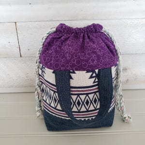 May include: A handmade drawstring bag with a purple top featuring a white circle pattern. The lower section is denim with a geometric design in white and burgundy. The bag has denim handles and a braided rope drawstring.