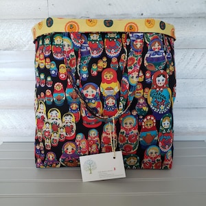 May include: A fabric tote bag featuring a repeating pattern of colorful Matryoshka dolls on a black background. The bag has a yellow floral trim and matching handles. A white tag with text hangs from the handle.