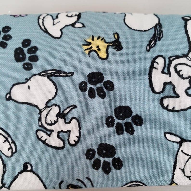 K&ouml;nnte beinhalten: Hellblauer Stoff mit einem sich wiederholenden Muster aus Snoopy, Woodstock und schwarzen Pfotenabdr&uuml;cken. Die wei&szlig;en Snoopy-Figuren sind in verschiedenen Posen, und Woodstock ist gelb. Der Stoff hat ein verspieltes, cartoonartiges Design.