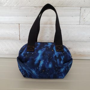 May include: A small, rounded tote bag with a celestial galaxy print in shades of blue and gold. The bag has black handles and a black zipper. The handles are attached with brown stitching. The bag is sitting on a white surface.