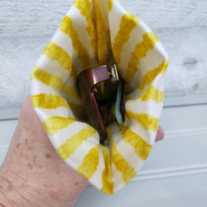 K&ouml;nnte beinhalten: Eine Brille in einem gelb-wei&szlig; gestreiften Etui. Die Brille hat einen ros&eacute;goldenen Rahmen und hellblaue B&uuml;gel. Das Etui ist aus Stoff.