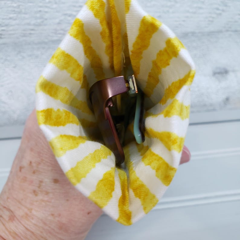 K&ouml;nnte beinhalten: Eine Brille in einem gelb-wei&szlig; gestreiften Etui. Die Brille hat einen ros&eacute;goldenen Rahmen und hellblaue B&uuml;gel. Das Etui ist aus Stoff.
