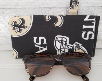 May include: Sunglasses with a tortoiseshell frame rest in front of a black eyeglass case. The case features a gold and black fleur-de-lis pattern and the word "SAINTS". A small tab is attached to the top of the case.