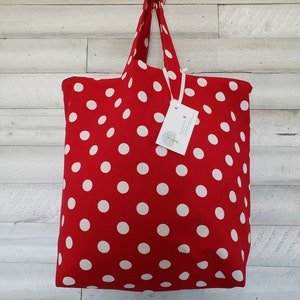 May include: Red tote bag with white polka dots. The bag is hanging on a hook against a white wood background. The bag has a white tag with the text "The Little Workshop" on it.