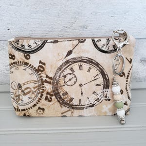 May include: A rectangular fabric zipper pouch featuring a beige background with a repeating dark brown vintage clock face pattern. A silver metal keychain is attached to the zipper pull, decorated with white, beige, and sage green silicone beads.