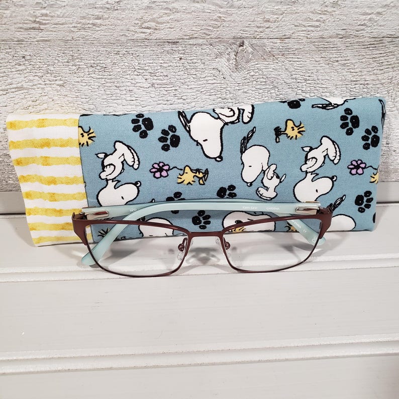 K&ouml;nnte beinhalten: Eine Brille mit braunem Metallrahmen und hellblauen Akzenten liegt auf einer wei&szlig;en Oberfl&auml;che. Ein blaues Brillenetui mit Snoopy-Muster und ein gelb-wei&szlig; gestreiftes Etui befinden sich hinter der Brille. Das Snoopy-Muster enth&auml;lt Pfotenabdr&uuml;cke und Blumen.
