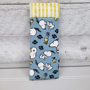K&ouml;nnte beinhalten: Ein Stoff-Brillenetui mit hellblauem Hintergrund, das Snoopy- und Woodstock-Figuren, Pfotenabdr&uuml;cke und Blumen zeigt. Der obere Teil hat gelbe und wei&szlig;e vertikale Streifen. Das Etui ist f&uuml;r Brillen oder Sonnenbrillen konzipiert.