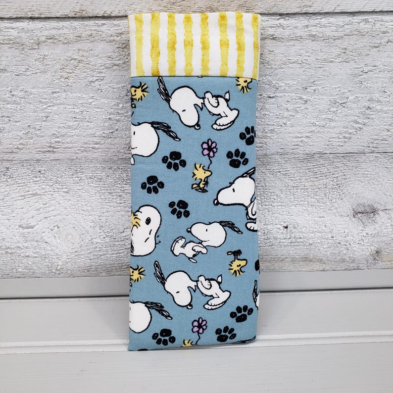 K&ouml;nnte beinhalten: Ein Stoff-Brillenetui mit hellblauem Hintergrund, das Snoopy- und Woodstock-Figuren, Pfotenabdr&uuml;cke und Blumen zeigt. Der obere Teil hat gelbe und wei&szlig;e vertikale Streifen. Das Etui ist f&uuml;r Brillen oder Sonnenbrillen konzipiert.