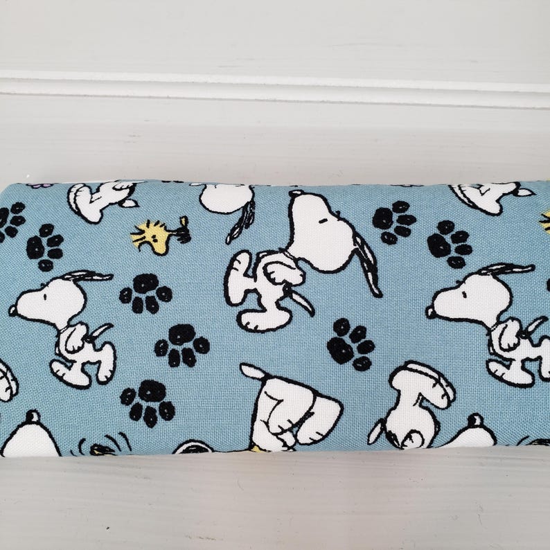 K&ouml;nnte beinhalten: Hellblaues Stoffmuster mit sich wiederholenden Snoopy- und schwarzen Pfotenabdruck-Motiven. Die Snoopy-Illustrationen sind wei&szlig; mit schwarzen Umrissen. Geeignet f&uuml;r N&auml;hprojekte.