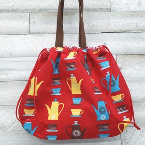 May include: A red drawstring bag with a repeating pattern of teapots and teacups in yellow, blue, and brown. The bag has brown handles and a drawstring closure. The bag is made of fabric.