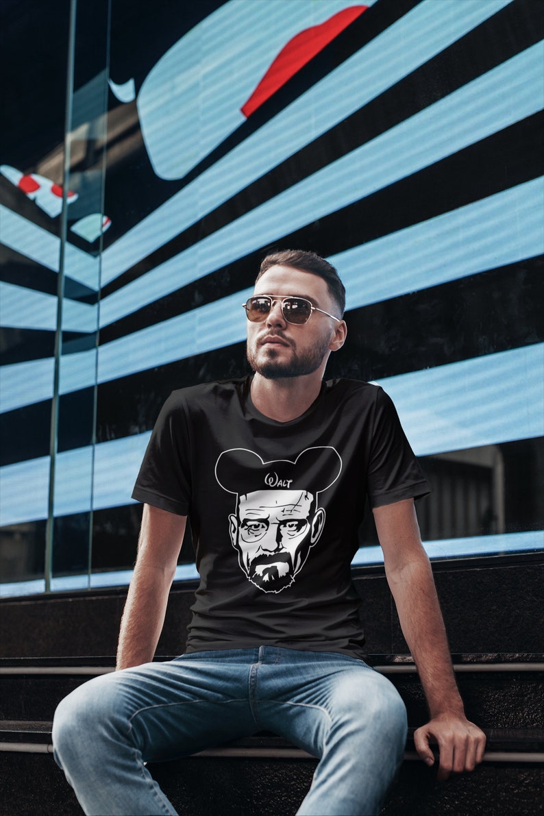 May include: A black t-shirt with a white graphic of Walter White from the TV show Breaking Bad wearing Mickey Mouse ears. The text "Walt" is written on the ears. The person wearing the shirt is looking to the right.