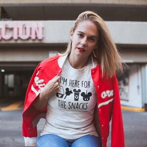 May include: A woman wearing a white t-shirt with the text "I'm here for the snacks" and black Mickey Mouse themed graphics. She is wearing a red bomber jacket with white sleeves and a white collar.