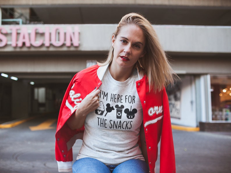 May include: A woman wearing a white t-shirt with the text "I'm here for the snacks" and black Mickey Mouse themed graphics. She is wearing a red bomber jacket with white sleeves and a white collar.