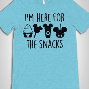 May include: A light blue t-shirt with the text "I'm here for the snacks" in black. The text is surrounded by black Mickey Mouse icons and images of snacks.