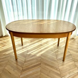 May include: Oval wooden dining table with four legs. The table has a light brown finish and a smooth surface. The legs are tapered and angled outwards. The table is approximately 152 cm long and 102 cm wide.