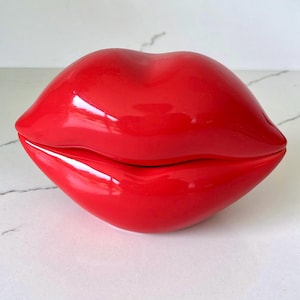 May include: A glossy, red lip-shaped decorative container. The container is a bright, vibrant red and has a smooth, reflective surface. The two halves of the lips are slightly parted, revealing a small opening. The container is a unique decorative item.