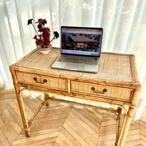 May include: A rectangular rattan desk featuring a woven top surface, two drawers with dark metal handles, and four sturdy legs. A silver laptop is open on the desk, and a small white bowl containing dark red flowers sits to the left of the laptop.