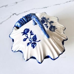 May include: A white ceramic basket with a blue handle and floral pattern. The basket features a scalloped edge and is adorned with blue roses and leaves. The handle is blue and has a textured design.