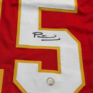 May include: Close-up of a red jersey with a large white number 5 outlined in gold. The number has a signature in black ink. A circular sticker with text is visible near the bottom of the number.