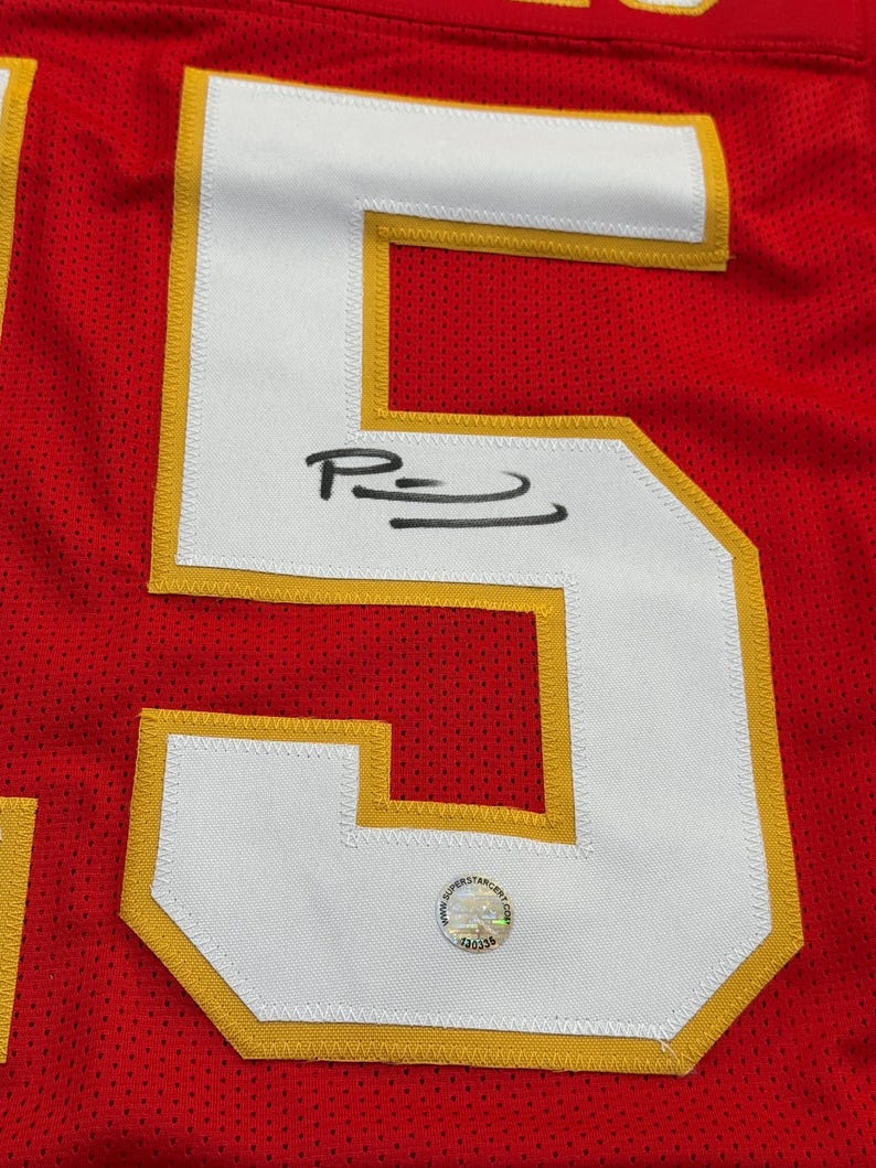 May include: Close-up of a red jersey with a large white number 5 outlined in gold. The number has a signature in black ink. A circular sticker with text is visible near the bottom of the number.