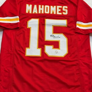 May include: Red football jersey with the name "MAHOMES" and the number "15" in white with gold trim. The jersey has a signature on the number. The sleeves have white and gold stripes.