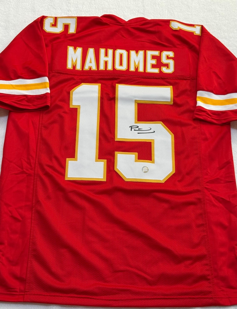 May include: Red football jersey with the name "MAHOMES" and the number "15" in white with gold trim. The jersey has a signature on the number. The sleeves have white and gold stripes.