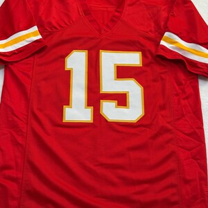 May include: A red football jersey with the number 15 in white with yellow trim on the front. The sleeves have white and yellow stripes. The jersey has a V-neck.