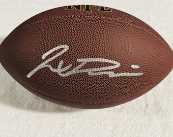 May include: A brown leather football with a textured surface featuring the NFL logo in gold and black at the top. A prominent silver autograph is written across the center of the ball, showcasing a fluid, stylized signature in a metallic finish.
