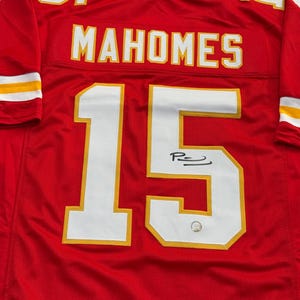 May include: Red football jersey with white and yellow accents. The jersey features the name "MAHOMES" and the number "15" in white. The jersey is signed with a black marker.
