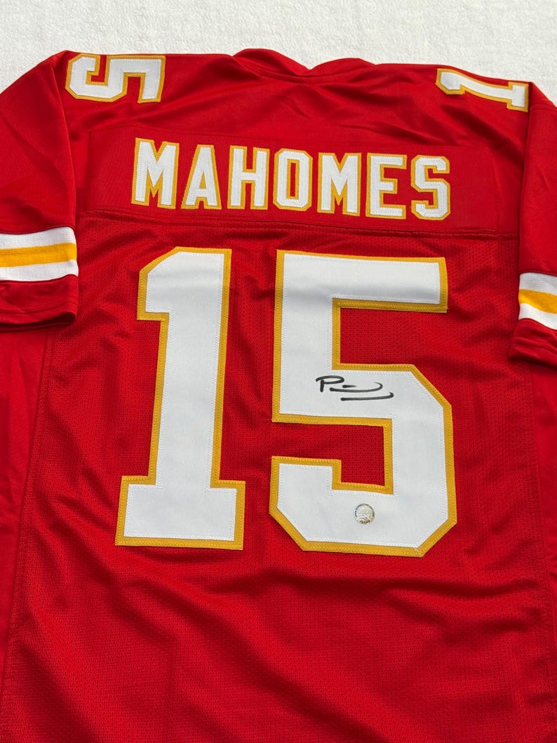 May include: Red football jersey with white and yellow accents. The jersey features the name "MAHOMES" and the number "15" in white. The jersey is signed with a black marker.