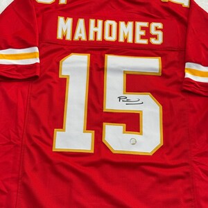 May include: Red football jersey with the name "MAHOMES" and the number "15" in white with gold trim. The jersey has a signature below the number. The sleeves have white and gold stripes.