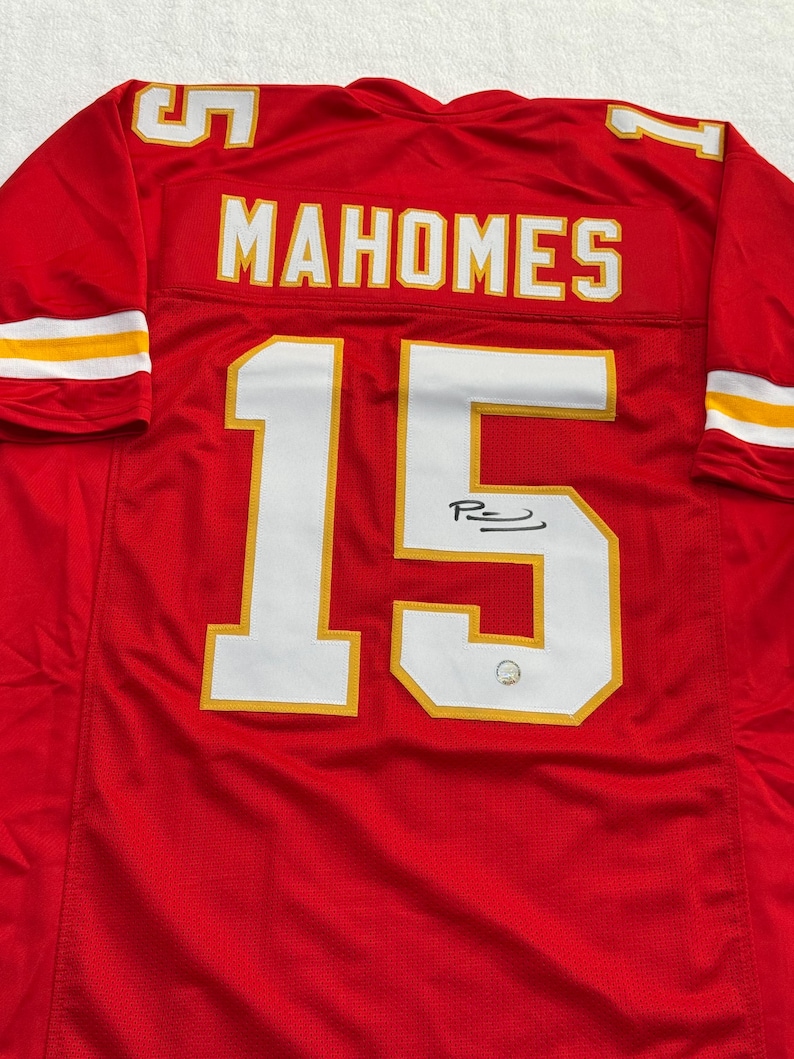 May include: Red football jersey with the name "MAHOMES" and the number "15" in white with gold trim. The jersey has a signature below the number. The sleeves have white and gold stripes.
