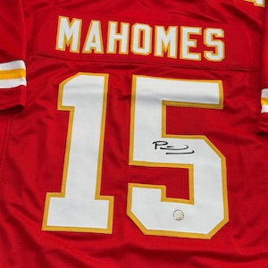 May include: A red football jersey with the name "MAHOMES" in white letters across the back and the number "15" in white with a gold outline. The jersey has a signature on the number 15.