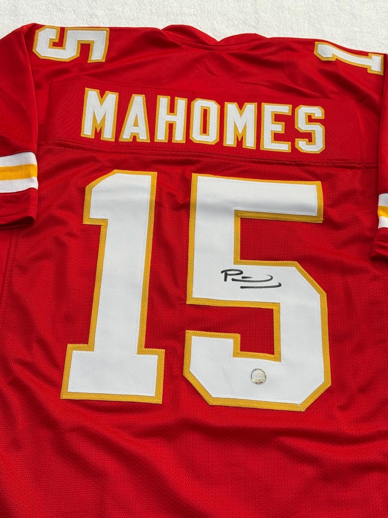 May include: A red football jersey with the name "MAHOMES" in white letters across the back and the number "15" in white with a gold outline. The jersey has a signature on the number 15.
