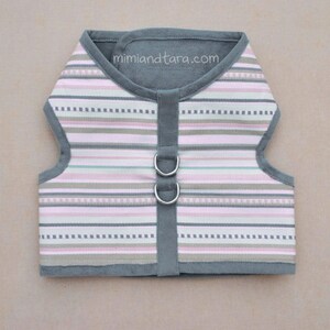 May include: A gray and pink striped dog harness with two silver D-rings. The harness has a gray fleece lining and the text "mimiandetara.com" is printed on the front.