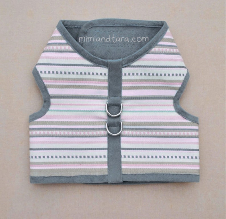 May include: A gray and pink striped dog harness with two silver D-rings. The harness has a gray fleece lining and the text "mimiandetara.com" is printed on the front.