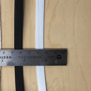 May include: Two pieces of elastic, one black and one white, are laid out next to a ruler. The ruler is marked in inches and shows that the elastic is 12 inches long.
