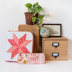 May include: A still-life arrangement featuring a quilted star-shaped case with a butterfly pattern, a pencil case with a whimsical print, and a small wooden drawer unit. A woven basket holds a potted plant, and a vintage globe and framed print complete the scene.