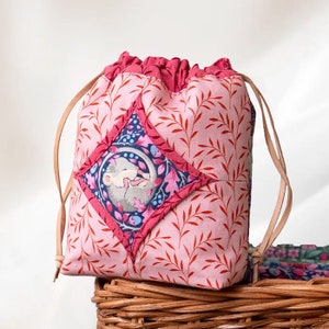 May include: A pink drawstring bag with a floral pattern and a diamond-shaped patch featuring a sleeping mouse. The bag has brown leather drawstrings.