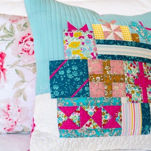 Puede incluir: Una almohada azul turquesa con un diseño de patchwork que presenta varios patrones florales y geométricos en rosa, amarillo, blanco y azul verdoso. La almohada está acolchada con un fondo blanco.