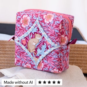 May include: A square fabric pouch featuring a vibrant pink and light blue floral pattern with a central diamond-shaped panel displaying a small tan squirrel. The bag includes a pink zipper closure and a small dark purple fabric tab on the side.